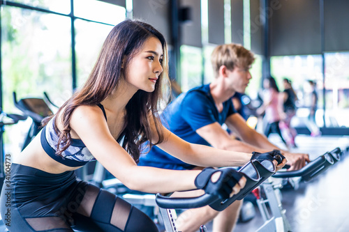 Young fit muscle Asian woman and Caucasian man exercising and working out in gym. People in sportswear cycling on exercise bike for cardio and focus on training for good health, wellness in fitness.
