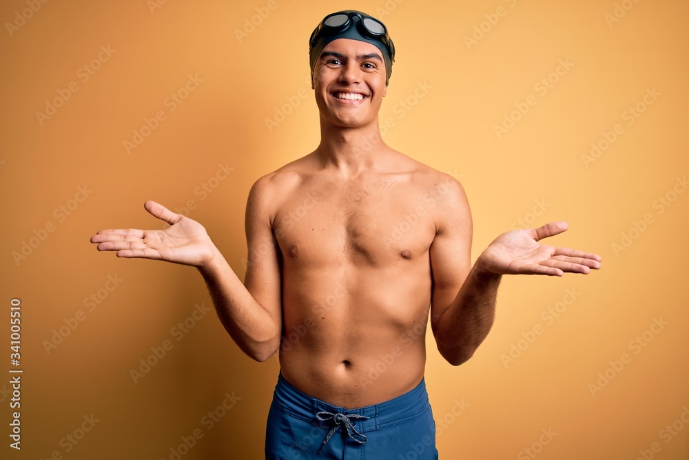 Young handsome man shirtless wearing swimsuit and swim cap over isolated yellow background smiling cheerful with open arms as friendly welcome, positive and confident greetings