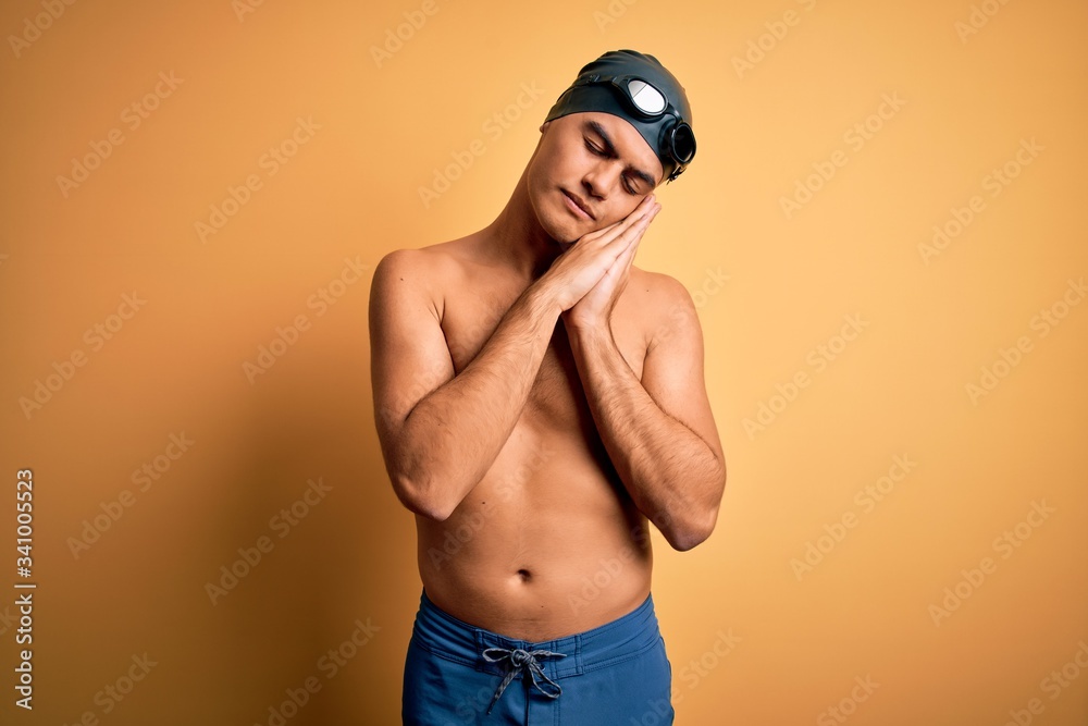 Young handsome man shirtless wearing swimsuit and swim cap over isolated yellow background sleeping tired dreaming and posing with hands together while smiling with closed eyes.