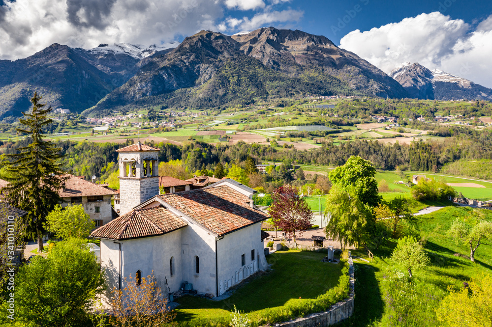 Aerial view of valley with church in Cares, Trentino, green slopes of the mountains of Italy, huge clouds over a valley, roof tops of houses, Dolomites on background