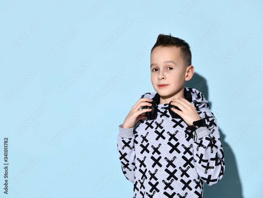 Teenage child with headphones on neck, in stylish hoodie and black bracelet. He smiling while posing against blue background Teenage child with headphones on neck, in stylish hoodie and black bracelet. He smiling while posing against blue background