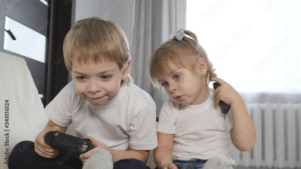 Blonde-haired children a girl and a boy play a game console. Happy children play sitting on a white sofa at home. Blonde-haired children a girl and a boy play a game console. Happy children play sitting on a white sofa at home.