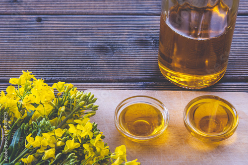 two glass cups with rapeseed oil and a glass bottle with oil near fresh rapeseed flowers