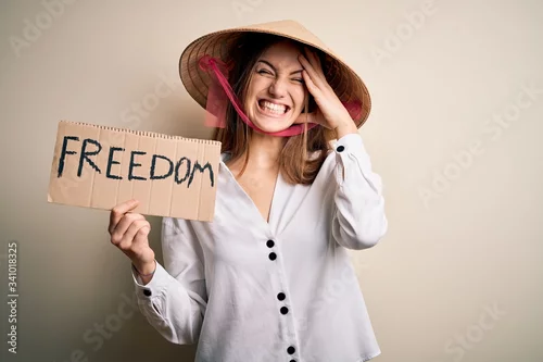 Obraz Young redhead woman wearing asian traditional hat holding banner with freedom message stressed with hand on head, shocked with shame and surprise face, angry and frustrated. Fear and upset