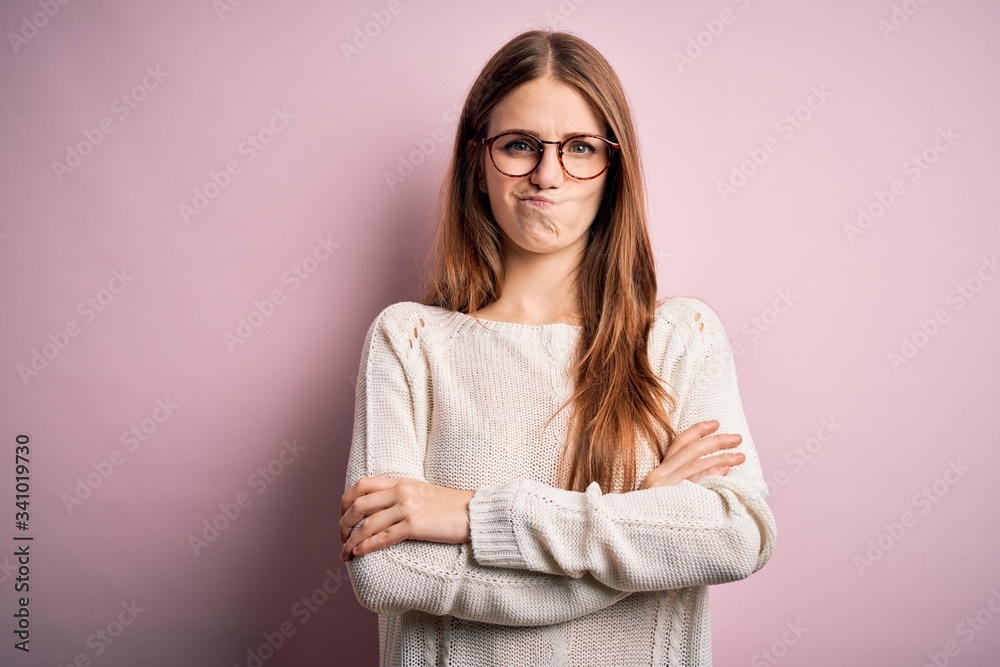 Young beautiful redhead woman wearing casual sweater and glasses over pink background skeptic and nervous, disapproving expression on face with crossed arms. Negative person. Young beautiful redhead woman wearing casual sweater and glasses over pink background skeptic and nervous, disapproving expression on face with crossed arms. Negative person.