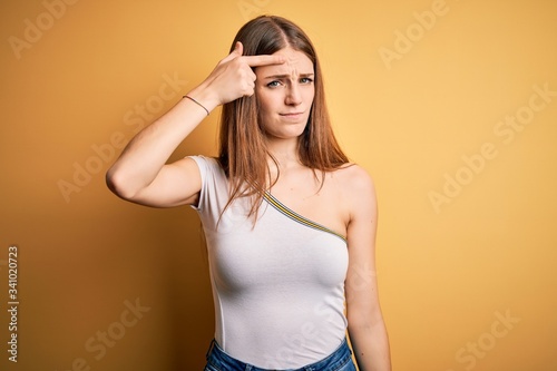 Young beautiful redhead woman wearing casual t-shirt over isolated yellow background pointing unhappy to pimple on forehead, ugly infection of blackhead. Acne and skin problem