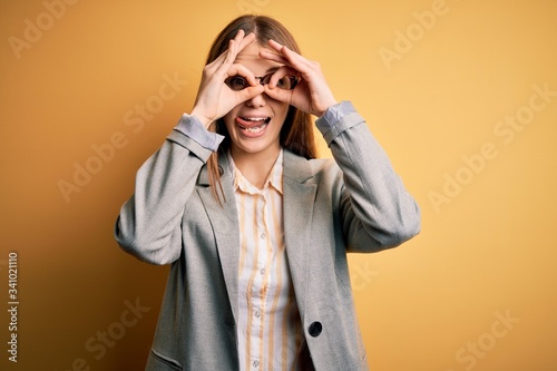 Young beautiful redhead woman wearing jacket and glasses over isolated yellow background doing ok gesture like binoculars sticking tongue out, eyes looking through fingers. Crazy expression.