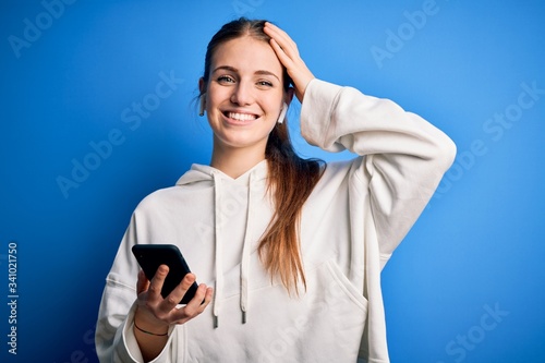 Young redhead sportswoman doing sport listening to music using earphones and smartphone smiling confident touching hair with hand up gesture, posing attractive and fashionable