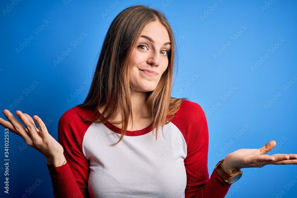 Fototapeta premium Young beautiful redhead woman wearing casual t-shirt over isolated blue background clueless and confused expression with arms and hands raised. Doubt concept.