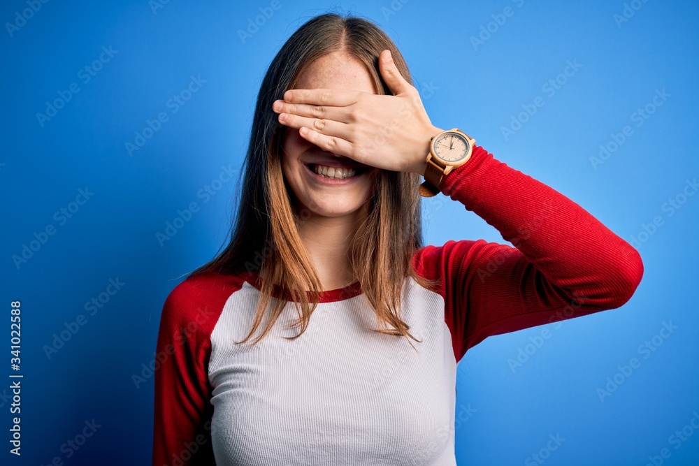 Fototapeta premium Young beautiful redhead woman wearing casual t-shirt over isolated blue background smiling and laughing with hand on face covering eyes for surprise. Blind concept.