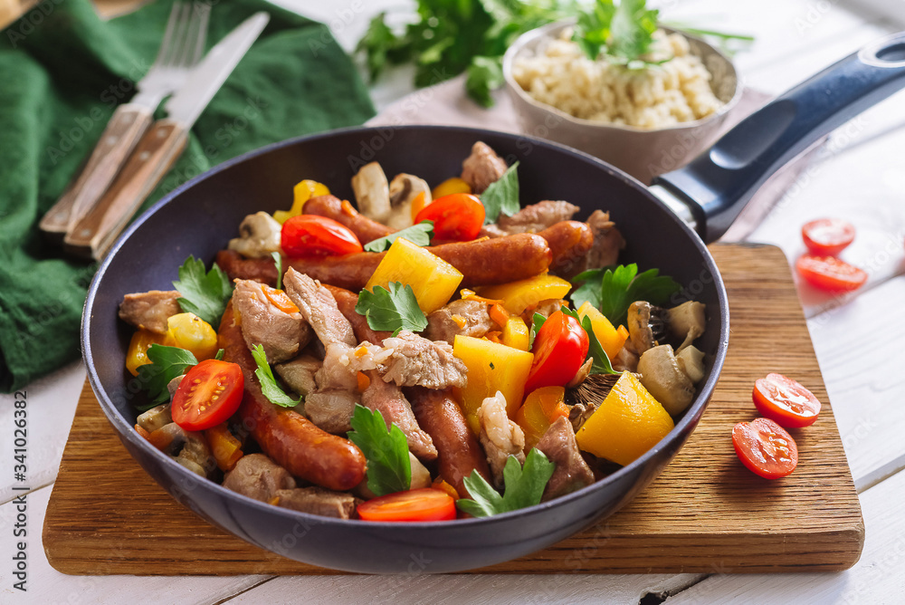 Frying pan with chopped pork meat, yellow bell pepper, cherry tomatoes, sausages, mushrooms, parsley leaves. A frying pan stands on a wooden board, stands for hot. Couscous grits in a bowl.