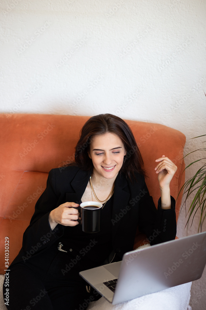 Fashionable young Caucasian woman sitting on sofa at home and working from home, remotely. She is keyboarding on laptop and drinking coffee. Freelance work concept. Copy space for text. Portrait. Fashionable young Caucasian woman sitting on sofa at home and working from home, remotely. She is keyboarding on laptop and drinking coffee. Freelance work concept. Copy space for text. Portrait.