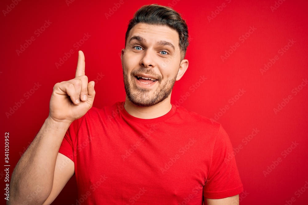 Fototapeta premium Young man with blue eyes wearing casual t-shirt over red isolated background pointing finger up with successful idea. Exited and happy. Number one.