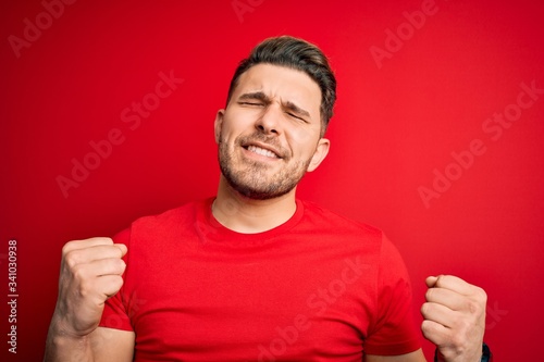 Young man with blue eyes wearing casual t-shirt over red isolated background very happy and excited doing winner gesture with arms raised, smiling and screaming for success. Celebration concept.