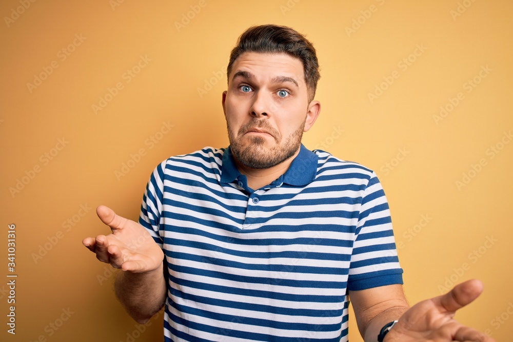 Young man with blue eyes wearing casual striped t-shirt over yellow background clueless and confused expression with arms and hands raised. Doubt concept. Young man with blue eyes wearing casual striped t-shirt over yellow background clueless and confused expression with arms and hands raised. Doubt concept.