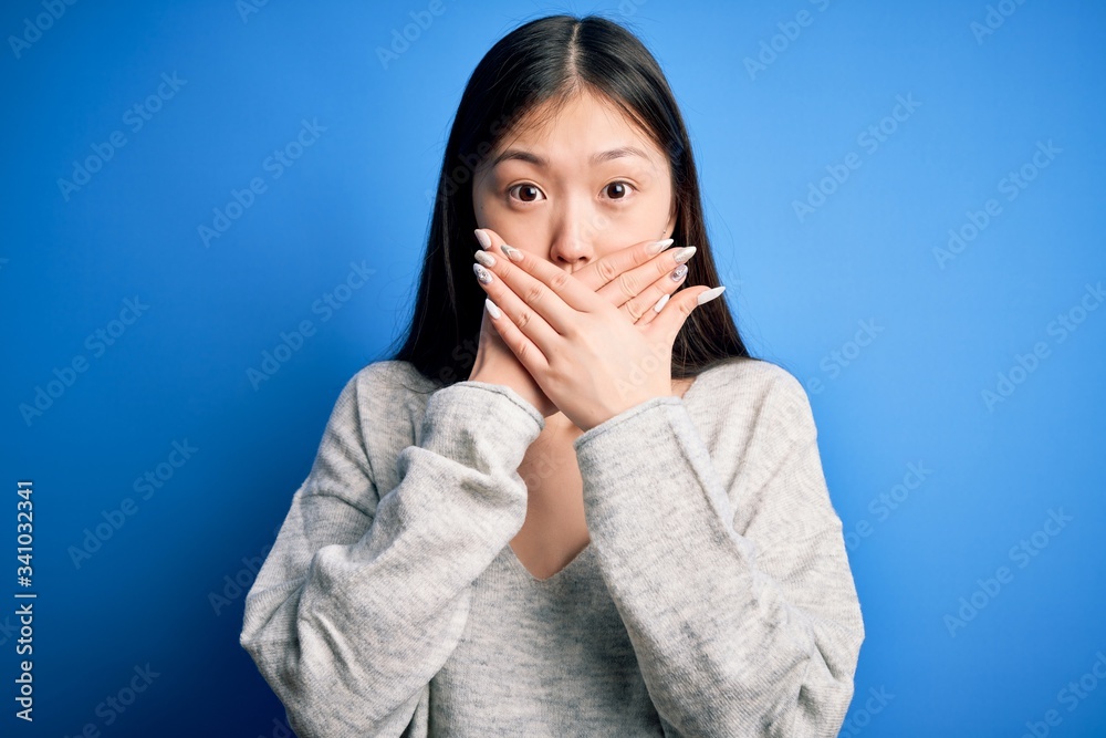 Young beautiful asian woman wearing casual sweater standing over blue isolated background shocked covering mouth with hands for mistake. Secret concept. Young beautiful asian woman wearing casual sweater standing over blue isolated background shocked covering mouth with hands for mistake. Secret concept.