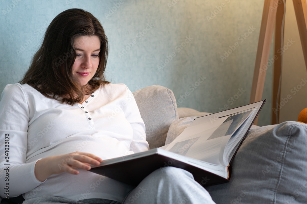 A pregnant young woman looking scrapbook photograph album and next to stands a carafe with water and fruit. A pregnant young woman looking scrapbook photograph album and next to stands a carafe with water and fruit.
