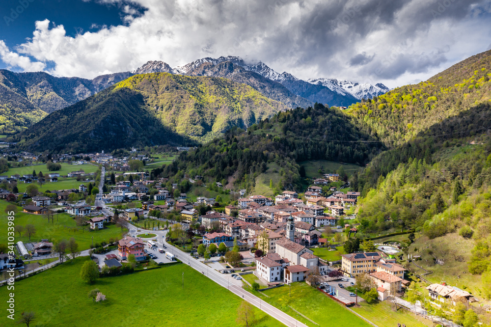 Aerial view of improbable green meadows of Italian Alps, Comano Terme, huge clouds over a valley, roof tops of houses, Dolomites on background, sunshines through clouds