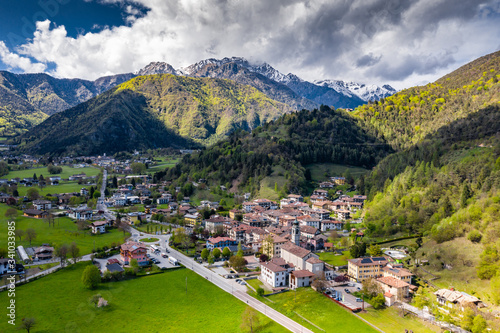 Aerial view of improbable green meadows of Italian Alps, Comano Terme, huge clouds over a valley, roof tops of houses, Dolomites on background, sunshines through clouds