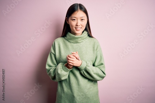 Young beautiful asian woman wearing green winter sweater over pink solated background with hands together and crossed fingers smiling relaxed and cheerful. Success and optimistic
