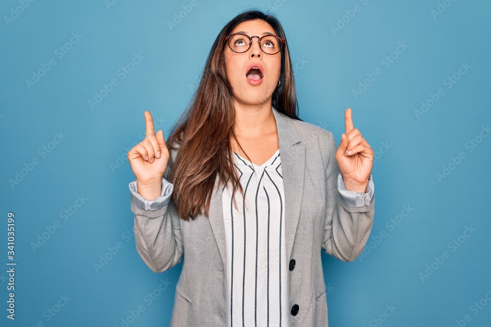 Young hispanic business woman wearing glasses standing over blue isolated background amazed and surprised looking up and pointing with fingers and raised arms. Young hispanic business woman wearing glasses standing over blue isolated background amazed and surprised looking up and pointing with fingers and raised arms.