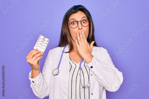 Young professional doctor woman holding pharmaceutical pills over purple background cover mouth with hand shocked with shame for mistake, expression of fear, scared in silence, secret concept