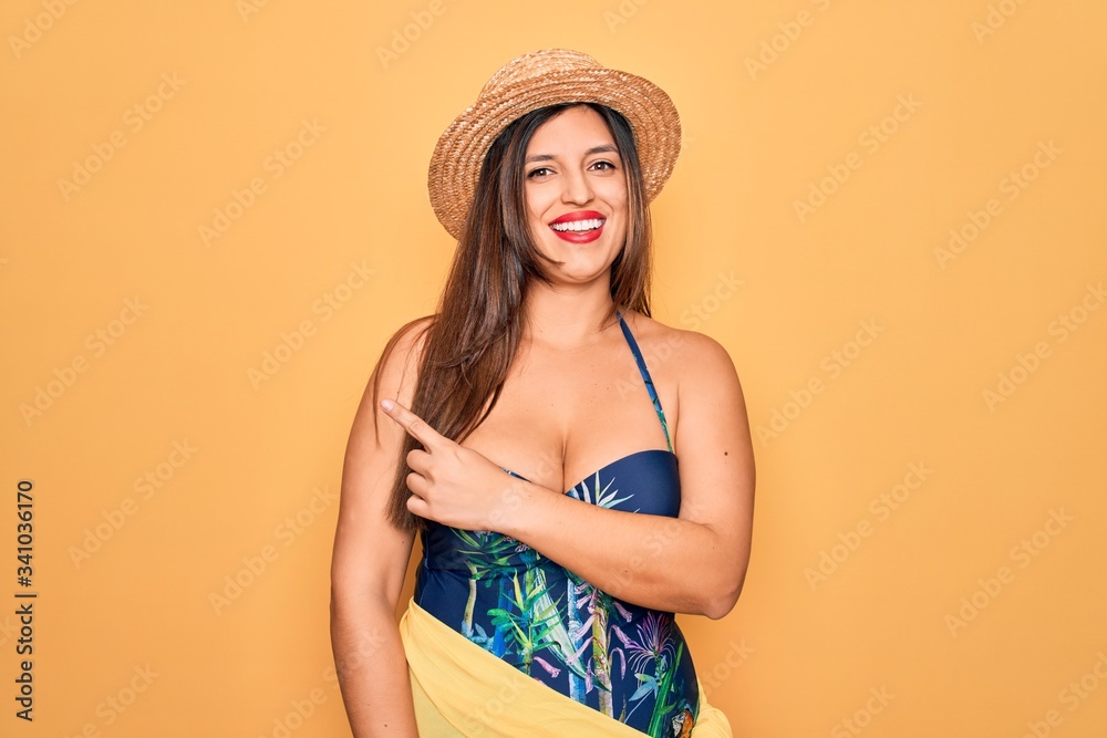 Young hispanic woman wearing summer hat and swimsuit over yellow background cheerful with a smile of face pointing with hand and finger up to the side with happy and natural expression on face Young hispanic woman wearing summer hat and swimsuit over yellow background cheerful with a smile of face pointing with hand and finger up to the side with happy and natural expression on face