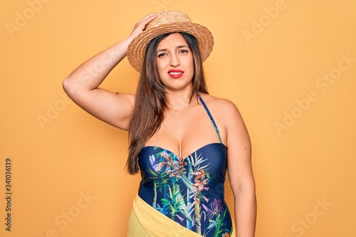 Young hispanic woman wearing summer hat and swimsuit over yellow background confuse and wonder about question. Uncertain with doubt, thinking with hand on head. Pensive concept.