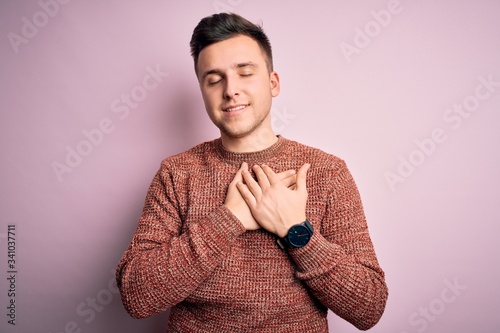 Young handsome caucasian man wearing casual winter sweater over pink isolated background smiling with hands on chest with closed eyes and grateful gesture on face. Health concept.