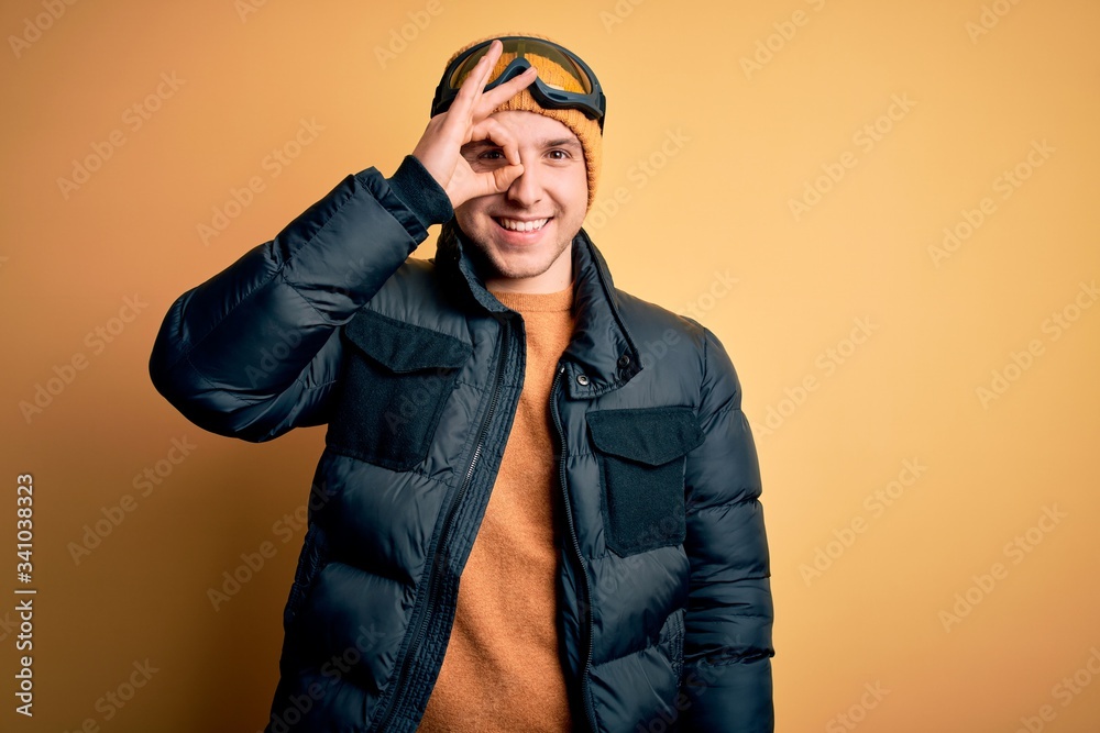 Young handsome caucasian man wearing hat, coat and ski glasses for winter and snow weather doing ok gesture with hand smiling, eye looking through fingers with happy face. Young handsome caucasian man wearing hat, coat and ski glasses for winter and snow weather doing ok gesture with hand smiling, eye looking through fingers with happy face.