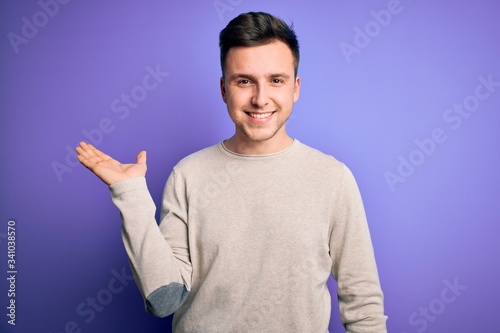 Young handsome caucasian man wearing casual sweater over purple isolated background smiling cheerful presenting and pointing with palm of hand looking at the camera.