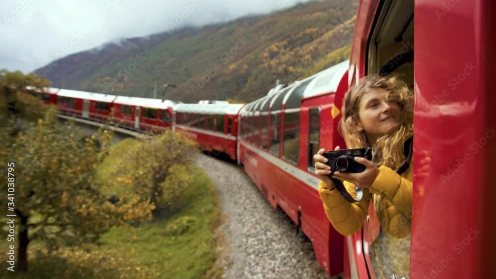 Inspiring female traveler and travel blogger look out of window of tourist train in cinematic beautiful mountains. Young beautiful woman hang out of window of red train. Excited world explorer