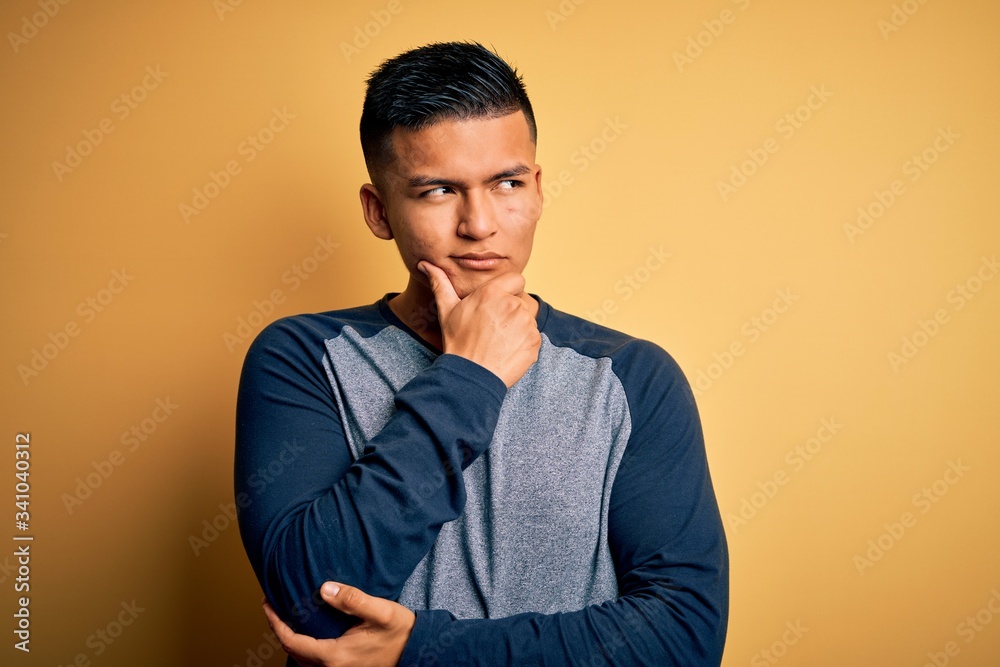 Young handsome latin man wearing casual t-shirt standing over yellow background with hand on chin thinking about question, pensive expression. Smiling with thoughtful face. Doubt concept. Young handsome latin man wearing casual t-shirt standing over yellow background with hand on chin thinking about question, pensive expression. Smiling with thoughtful face. Doubt concept.