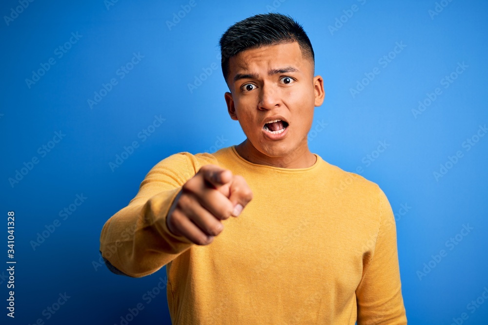Young handsome latin man wearing yellow casual sweater over isolated blue background pointing displeased and frustrated to the camera, angry and furious with you Young handsome latin man wearing yellow casual sweater over isolated blue background pointing displeased and frustrated to the camera, angry and furious with you