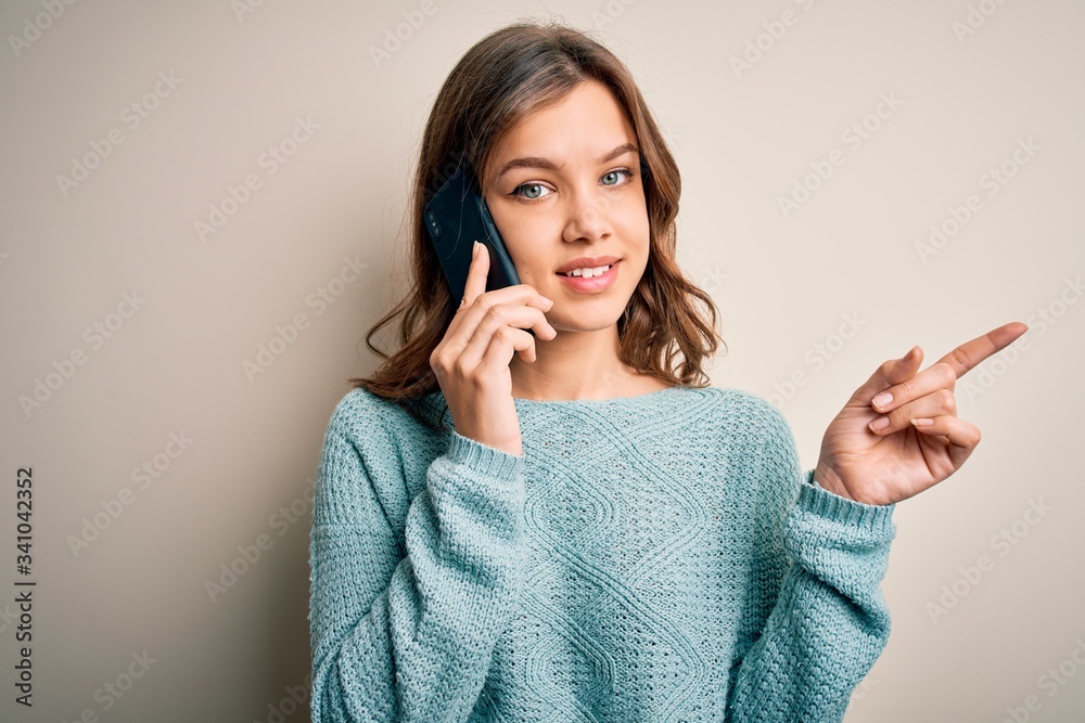 Young blonde girl having a conversation talking on smartphone over isolated background very happy pointing with hand and finger to the side Young blonde girl having a conversation talking on smartphone over isolated background very happy pointing with hand and finger to the side