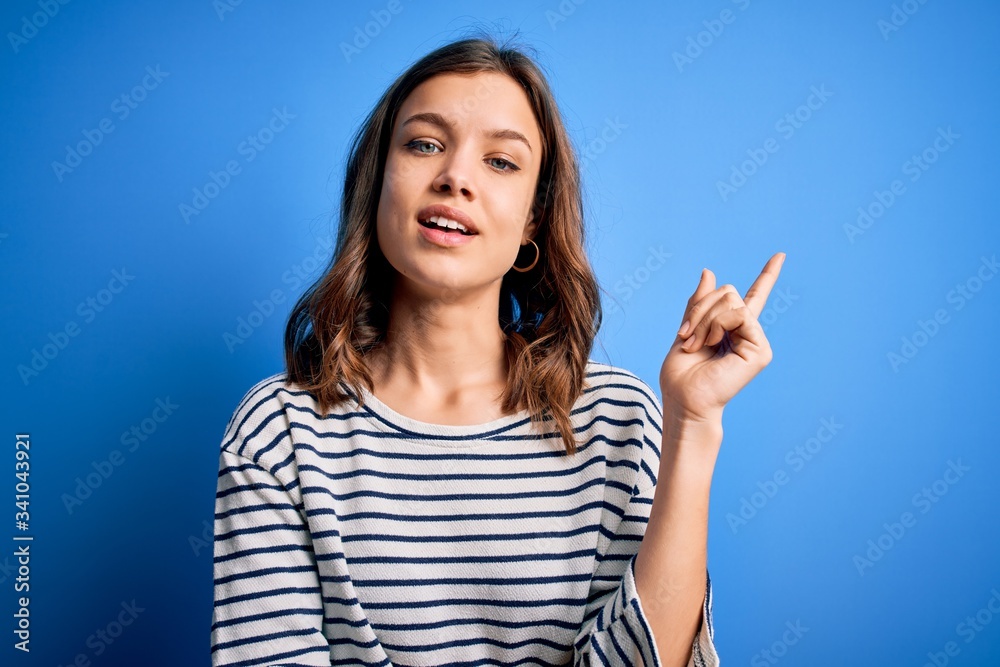 Young beautiful blonde girl wearing casual sweater standing over blue isolated background with a big smile on face, pointing with hand and finger to the side looking at the camera. Young beautiful blonde girl wearing casual sweater standing over blue isolated background with a big smile on face, pointing with hand and finger to the side looking at the camera.