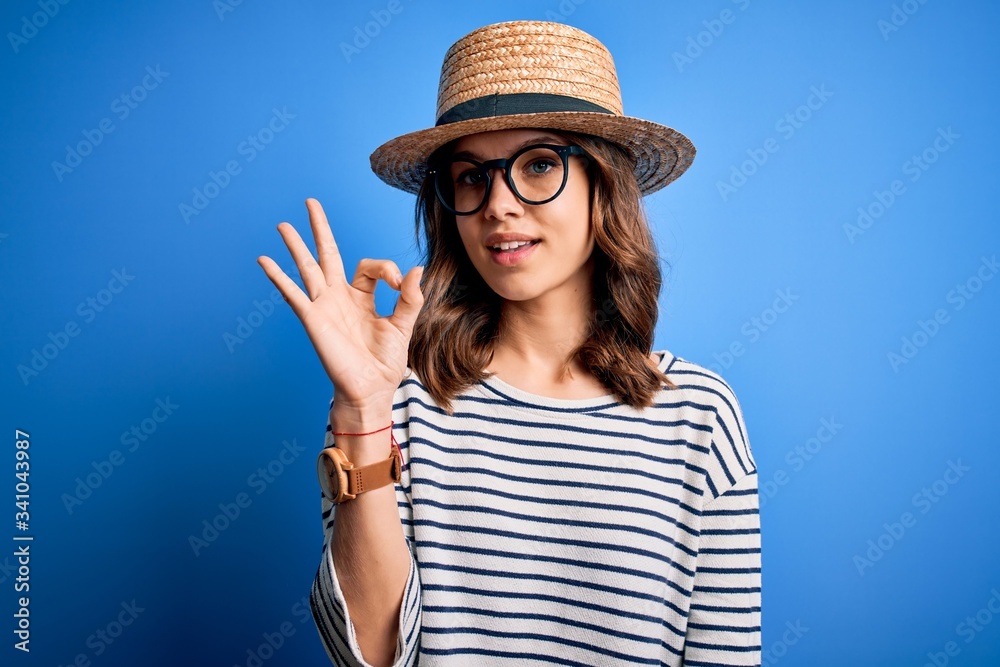 Young beautiful blonde girl wearing glasses and hat over blue isolated background doing ok sign with fingers, excellent symbol Young beautiful blonde girl wearing glasses and hat over blue isolated background doing ok sign with fingers, excellent symbol