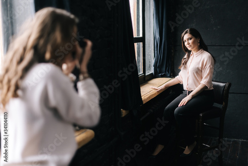 Woman model posing while sitting on chair in photo studio for unrecognizable female photographer which shooting with camera. Concept of creative work in photo studio, backstage job.