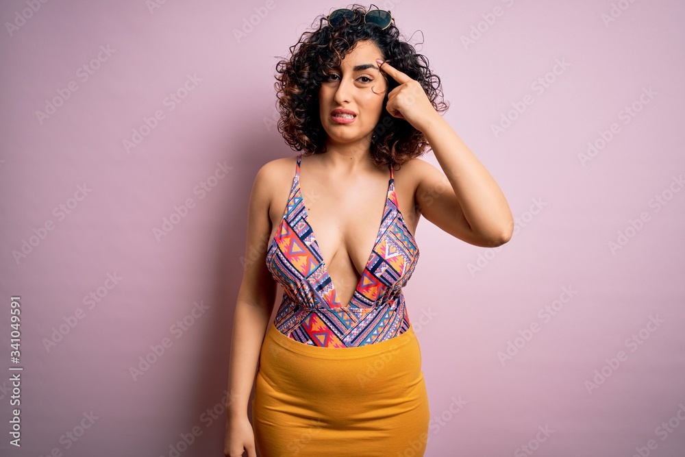 Young beautiful arab woman on vacation wearing swimsuit and sunglasses over pink background pointing unhappy to pimple on forehead, ugly infection of blackhead. Acne and skin problem Young beautiful arab woman on vacation wearing swimsuit and sunglasses over pink background pointing unhappy to pimple on forehead, ugly infection of blackhead. Acne and skin problem