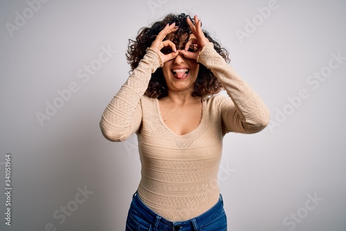 Young beautiful curly arab woman wearing casual t-shirt and glasses over white background doing ok gesture like binoculars sticking tongue out, eyes looking through fingers. Crazy expression.