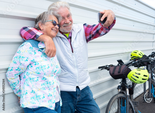 Two senior adult people stop riding an electric bicycle and rest against a white wall taking a selfie with the cell phones. A smiling couple with casual clothes