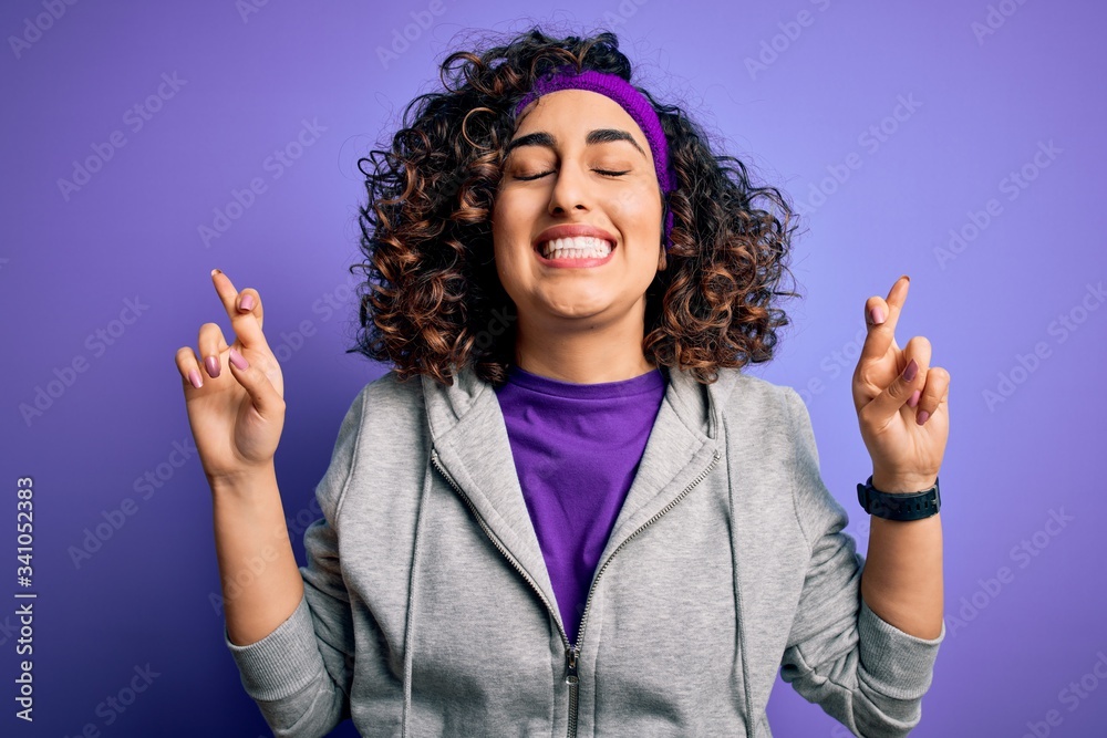 Beautiful curly arab sportswoman doing sport wearing sportswear over purple background gesturing finger crossed smiling with hope and eyes closed. Luck and superstitious concept. Beautiful curly arab sportswoman doing sport wearing sportswear over purple background gesturing finger crossed smiling with hope and eyes closed. Luck and superstitious concept.