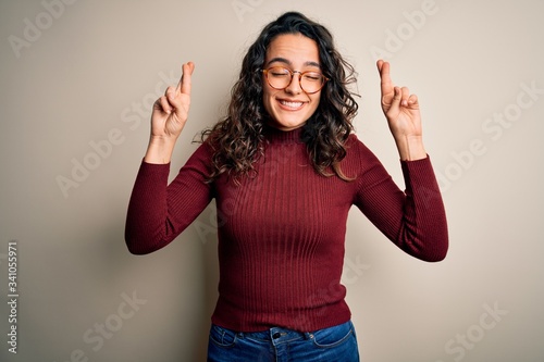 Beautiful woman with curly hair wearing casual sweater and glasses over white background gesturing finger crossed smiling with hope and eyes closed. Luck and superstitious concept.