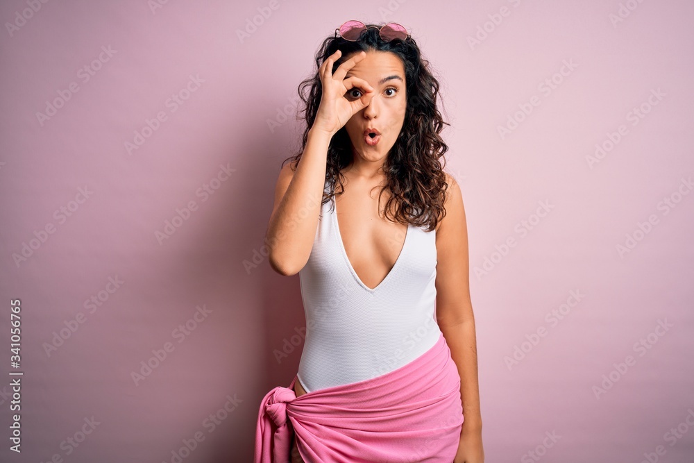 Beautiful woman with curly hair on vacation wearing white swimsuit over pink background doing ok gesture shocked with surprised face, eye looking through fingers. Unbelieving expression. Beautiful woman with curly hair on vacation wearing white swimsuit over pink background doing ok gesture shocked with surprised face, eye looking through fingers. Unbelieving expression.