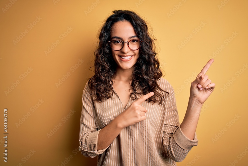 Beautiful woman with curly hair wearing striped shirt and glasses over yellow background smiling and looking at the camera pointing with two hands and fingers to the side. Beautiful woman with curly hair wearing striped shirt and glasses over yellow background smiling and looking at the camera pointing with two hands and fingers to the side.