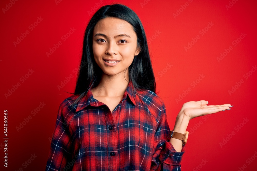 Young beautiful chinese woman wearing casual shirt over isolated red background smiling cheerful presenting and pointing with palm of hand looking at the camera.