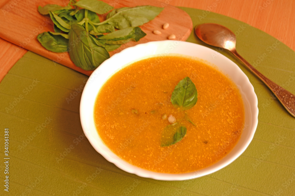 Top view of a plate of pumpkin soup with basil leaves and pine nuts on wooden plate and green background with a spoon Top view of a plate of pumpkin soup with basil leaves and pine nuts on wooden plate and green background with a spoon