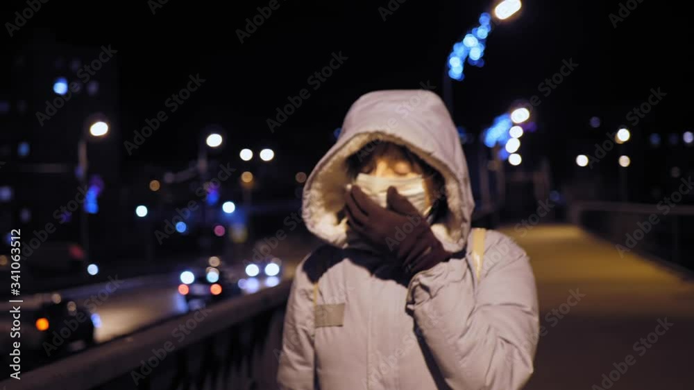 Young woman in a protective face mask coughs while hiding with a hand in a brown glove. A female patient with severe pain stops at the railing of the bridge late in the evening against a busy road. Young woman in a protective face mask coughs while hiding with a hand in a brown glove. A female patient with severe pain stops at the railing of the bridge late in the evening against a busy road.