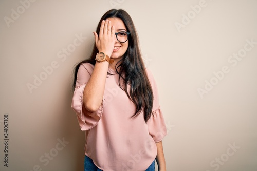 Young brunette elegant woman wearing glasses over isolated background covering one eye with hand, confident smile on face and surprise emotion.