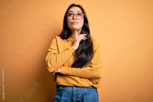 Young brunette woman wearing glasses and casual sweater over yellow isolated background Thinking concentrated about doubt with finger on chin and looking up wondering © Krakenimages.com